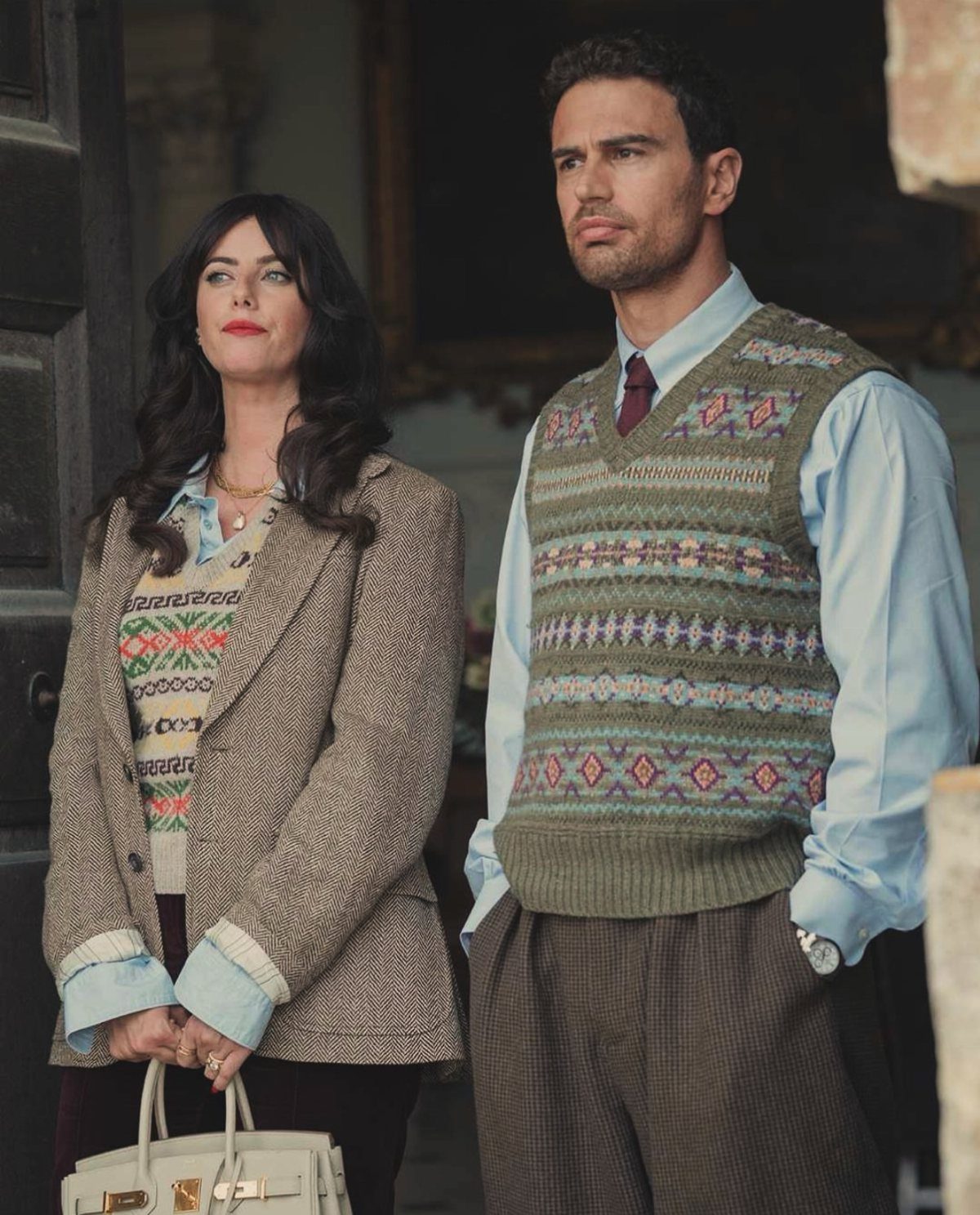 A man wearing a patterned olive green knit vest and a woman in a tweed blazer.
