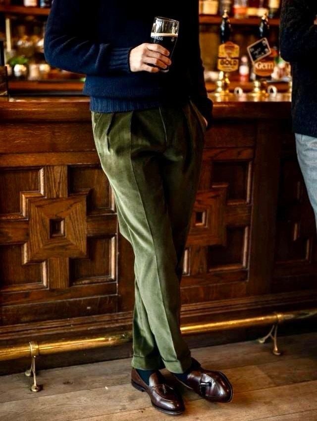 A man wearing olive corduroy trousers and leather loafers standing in a traditional wooden pub.