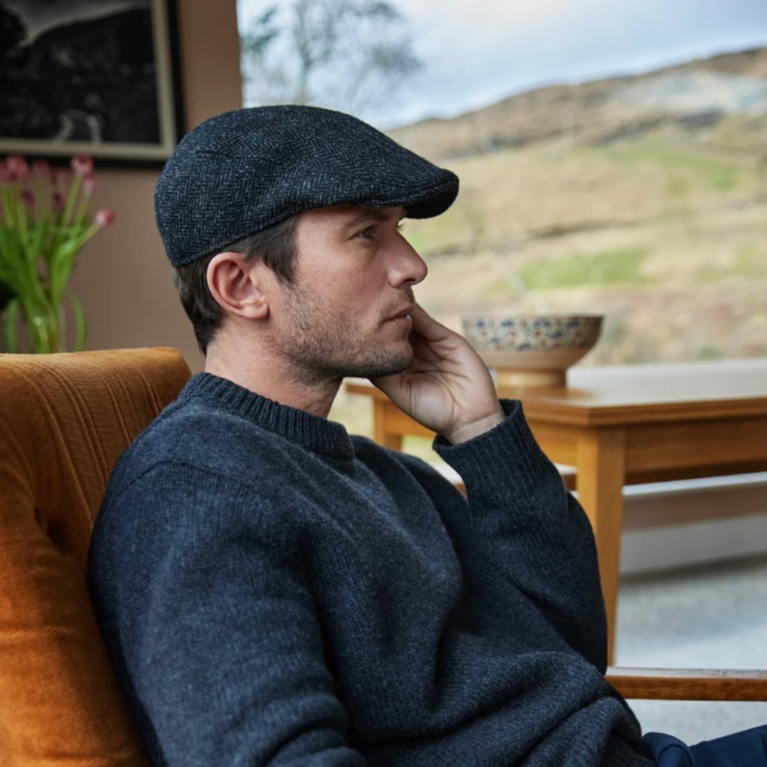 A man wearing a dark grey textured flat cap and a charcoal sweater sitting outdoors.