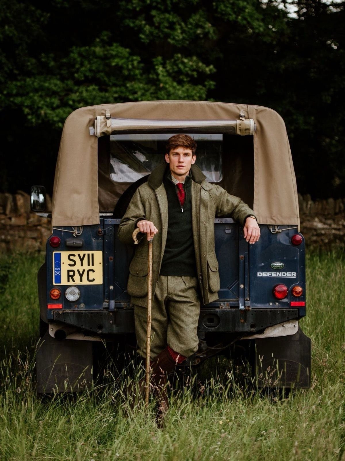 Man in layered country attire standing by a Land Rover in a grassy field.