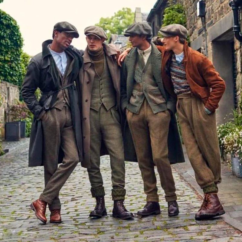 Four men posing in cobblestone street wearing various British country style clothing and flat caps.