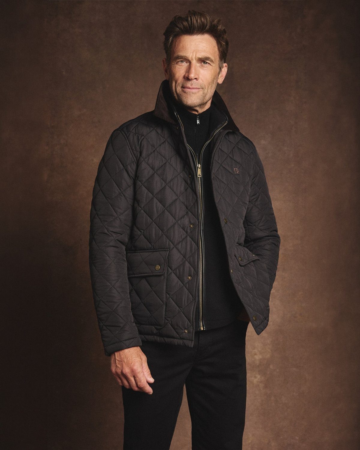 A man wearing a black diamond-quilted jacket and black turtleneck against a brown background.