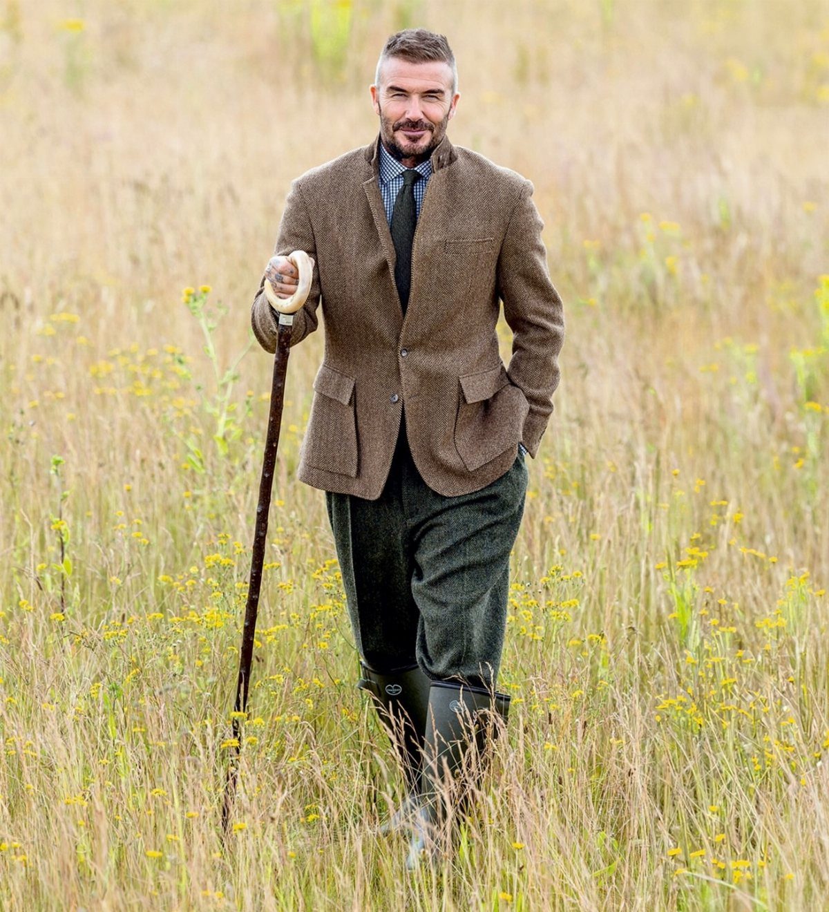 A man in traditional tweed country attire walking through a grassy field with a walking stick.
