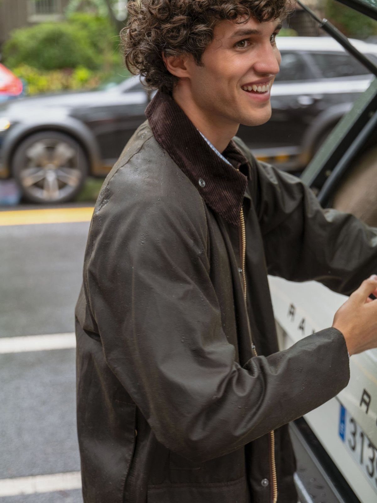 A smiling young man with curly hair wearing a dark olive green waxed jacket.