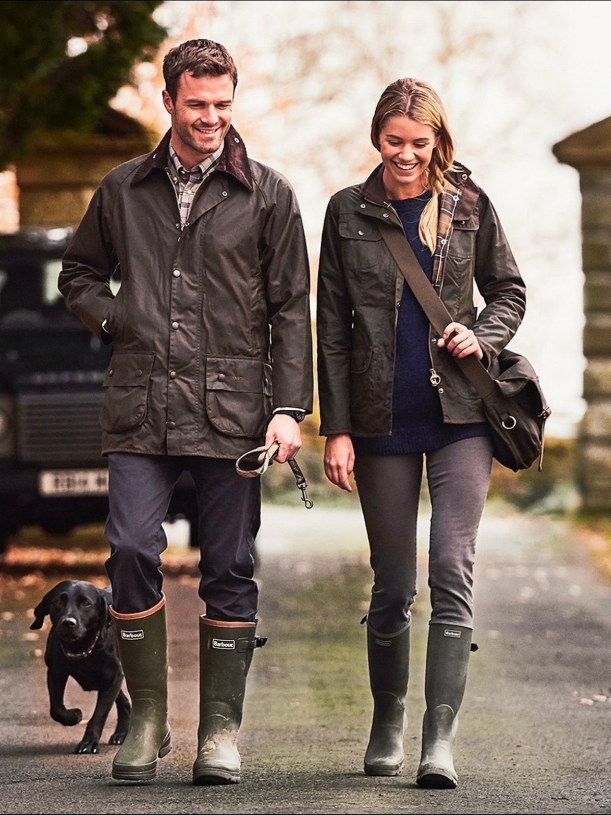 A man and woman walking a black dog while wearing waxed jackets and rubber wellington boots.