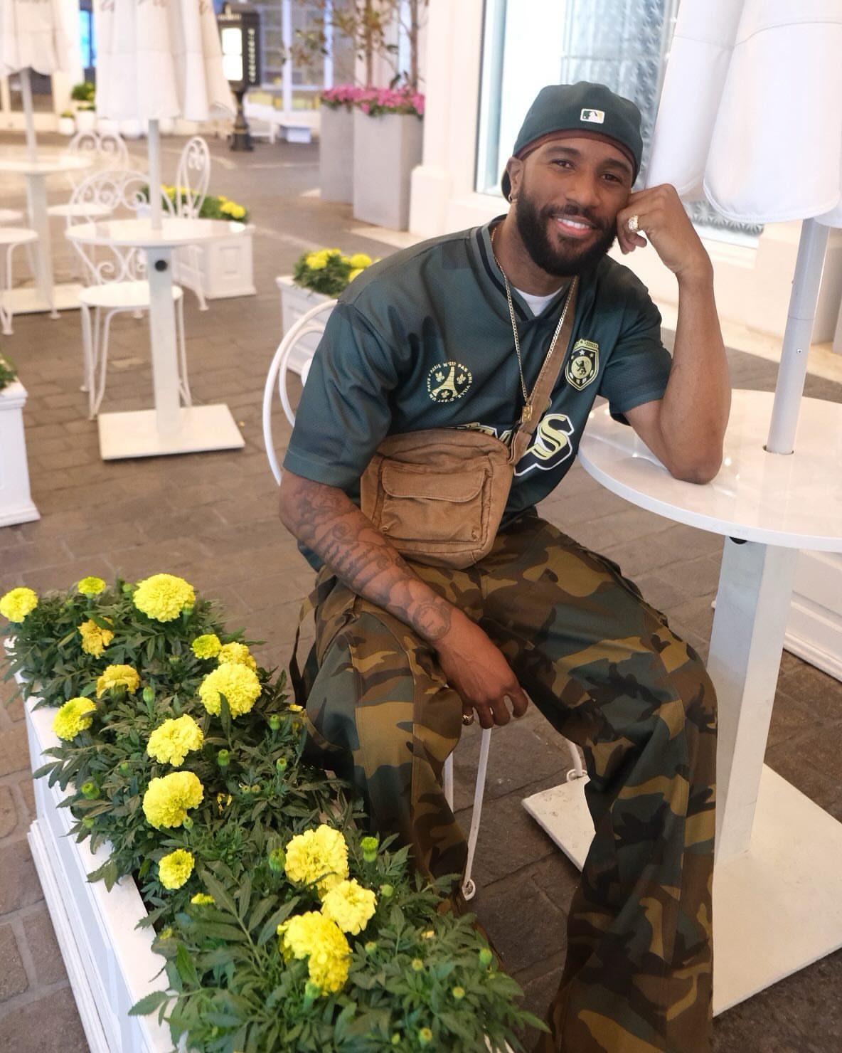 Man in camouflage cargo pants and a tan crossbody bag sitting at an outdoor cafe table.