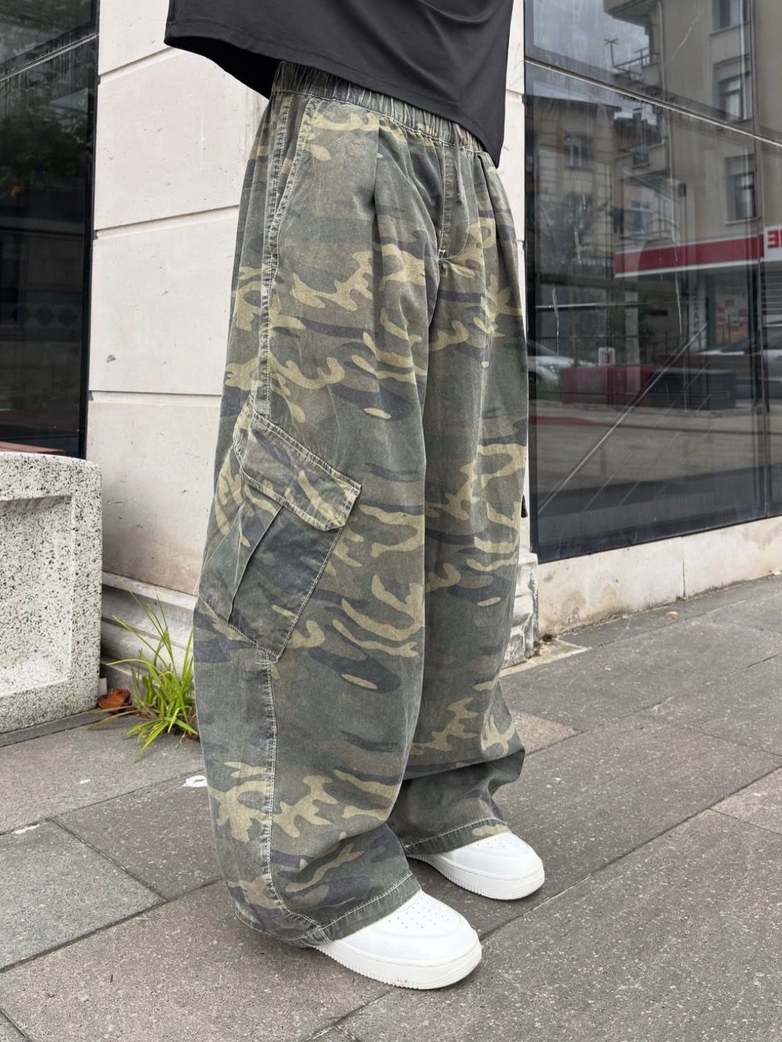 Person wearing camouflage cargo pants and white sneakers standing on a city sidewalk.