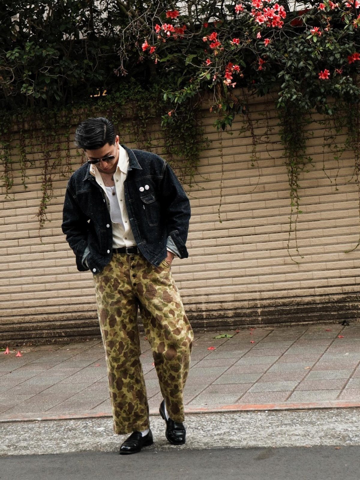 Man wearing a dark denim jacket, white shirt, and camouflage cargo pants standing by a brick wall.