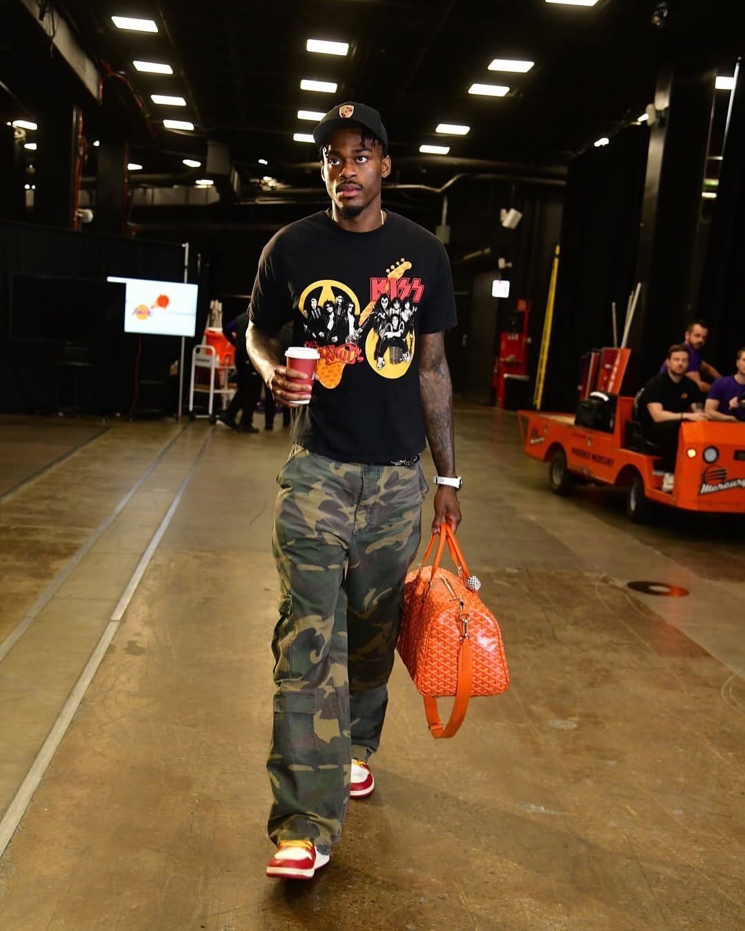 Man walking in camouflage cargo pants and a black graphic t-shirt while carrying an orange bag.