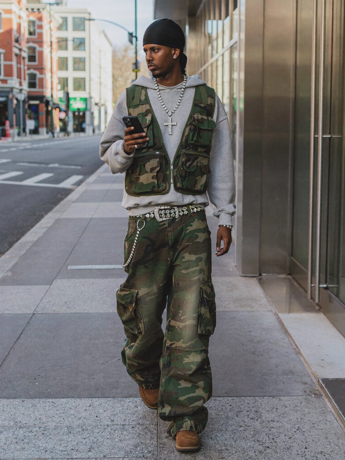 Man in camouflage cargo pants, grey hoodie, and utility vest walking on a city sidewalk.