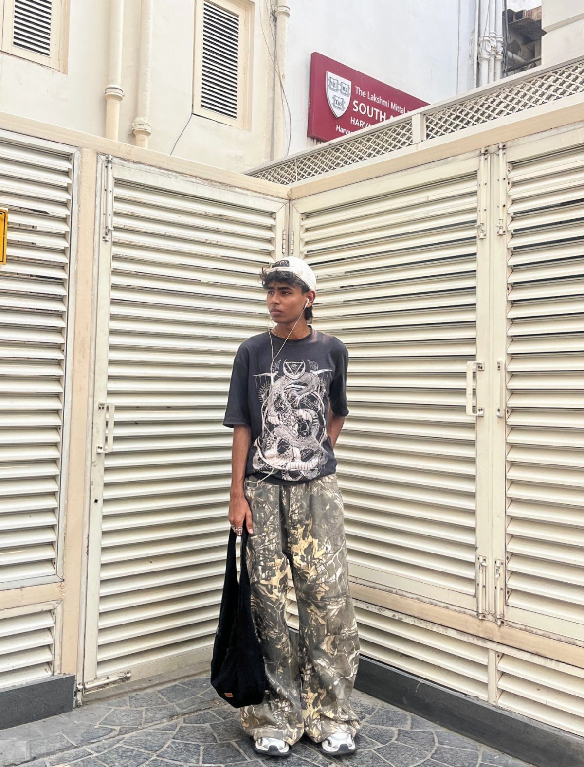 A person wearing a graphic tee and camouflage pants standing in front of white slatted doors.