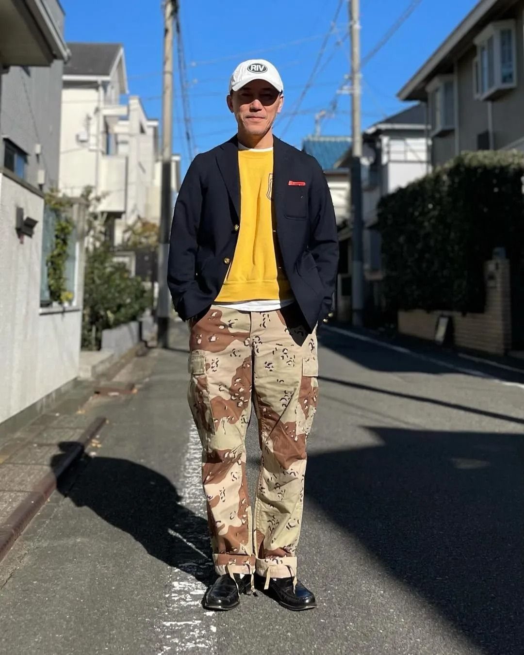 A man wearing tan camo pants, a yellow shirt, navy blazer, and a white baseball cap.
