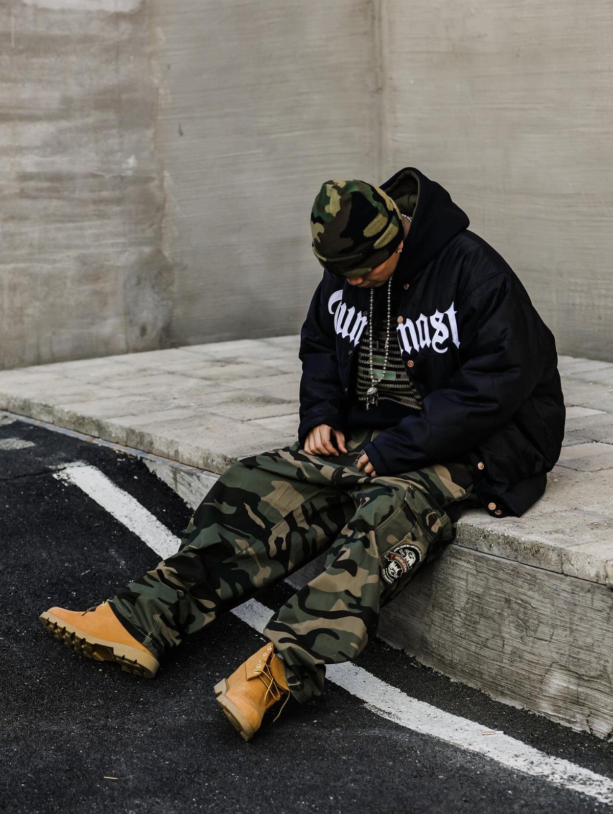 A person wearing camouflage pants, a black jacket, and tan boots sitting on a concrete ledge.
