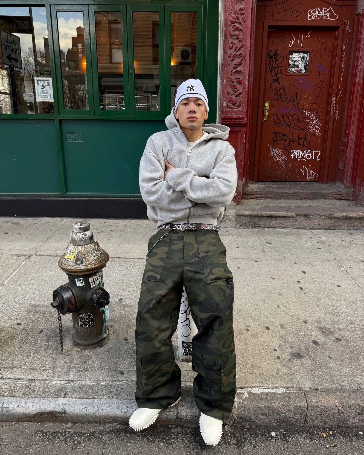 A person in camouflage pants and a grey cropped hoodie standing on a city sidewalk.