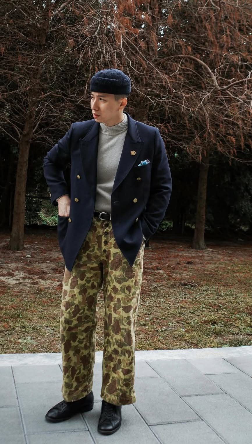 A person wearing camouflage pants, a grey turtleneck, and a navy oversized blazer standing outdoors.