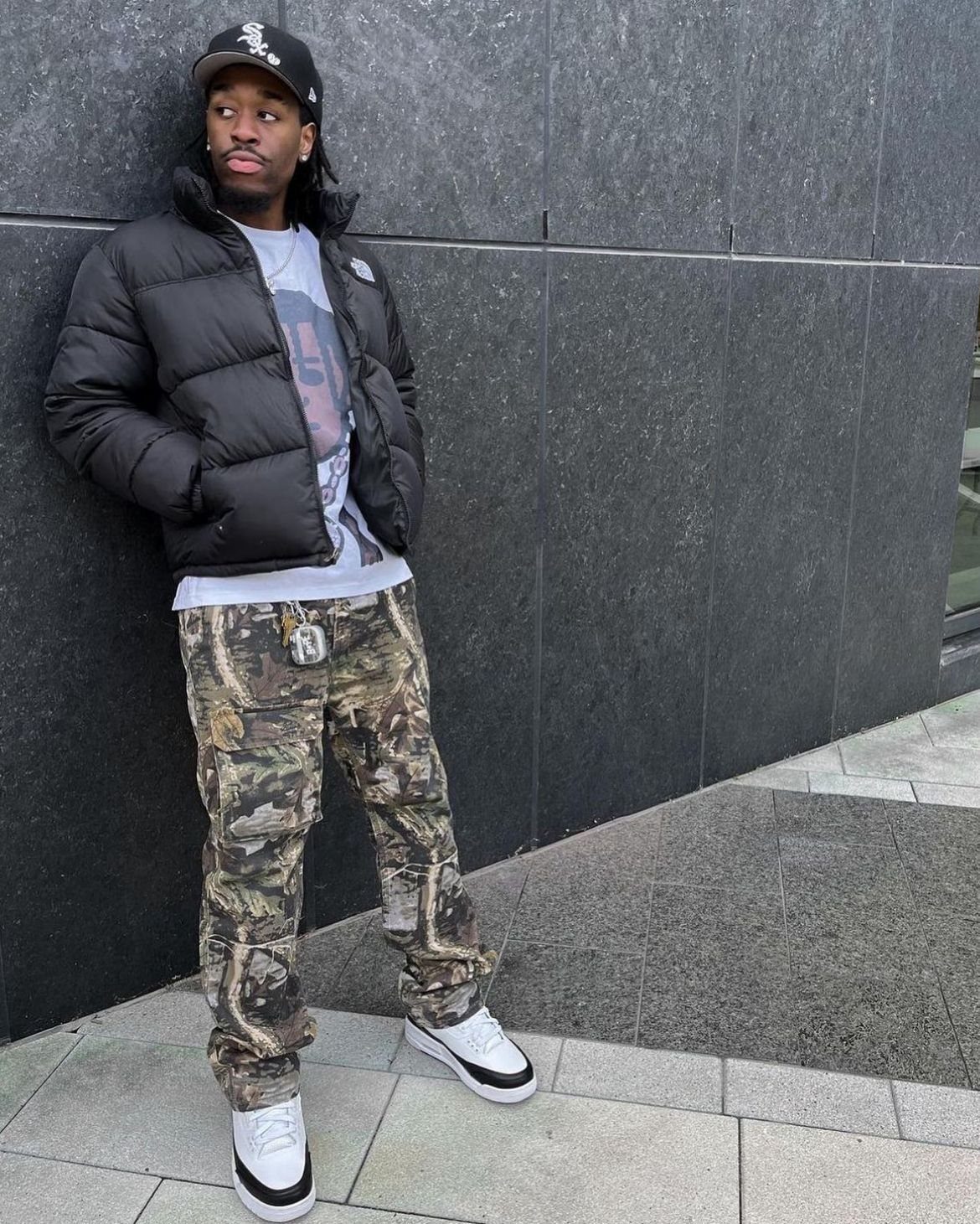 Man in black puffer jacket, graphic tee, and camo pants leaning against a wall.