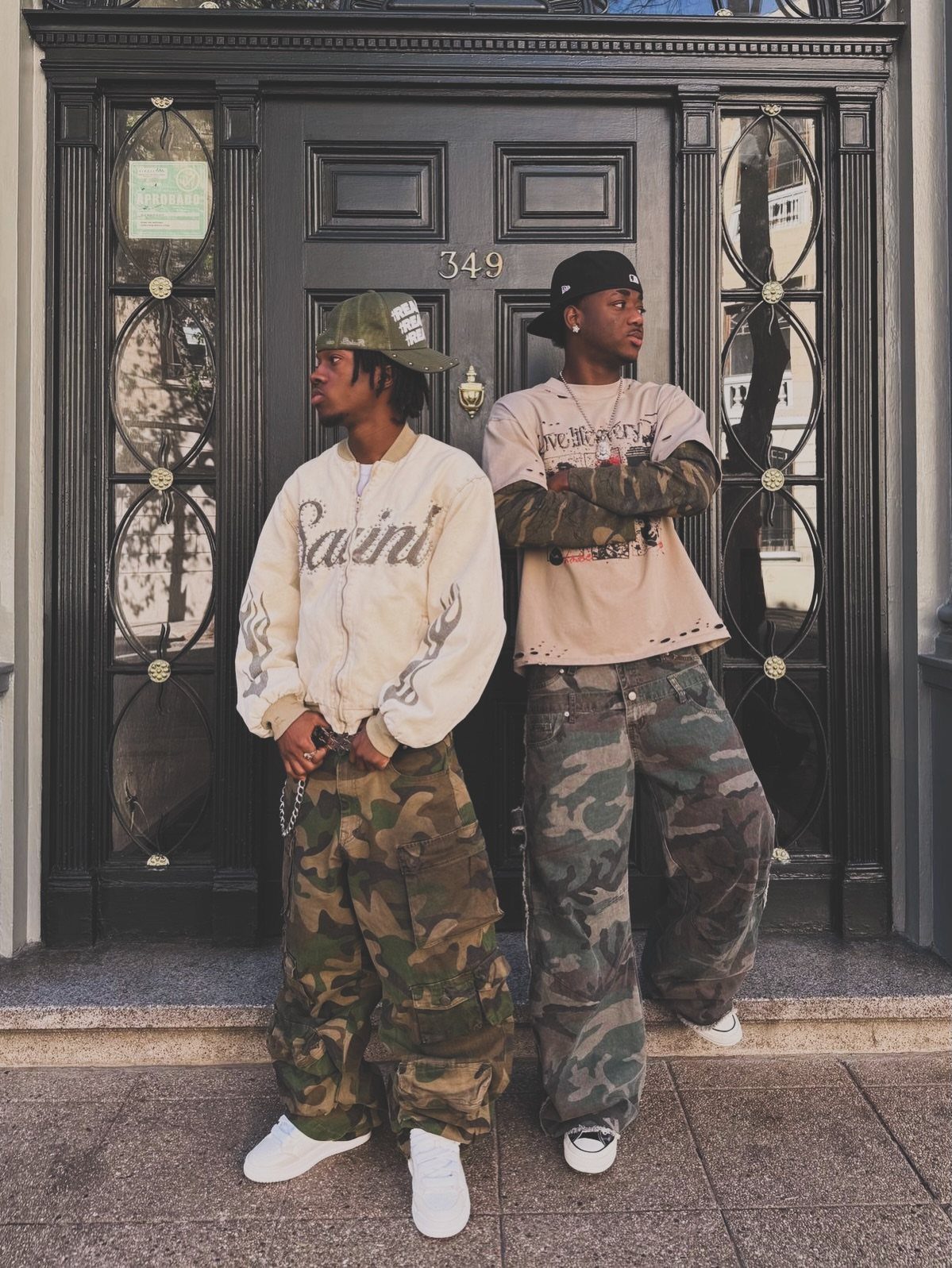 Two men posing in camouflage pants, streetwear tops, and white sneakers in front of a dark door.