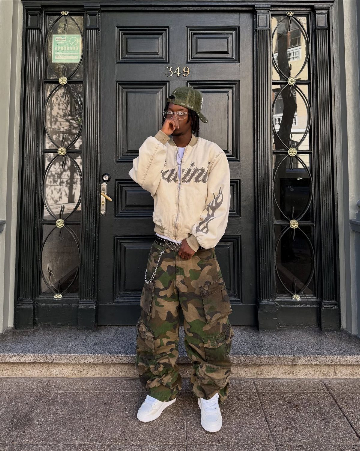 Person wearing camouflage cargo pants, a cream bomber jacket, and white sneakers standing before a black door.