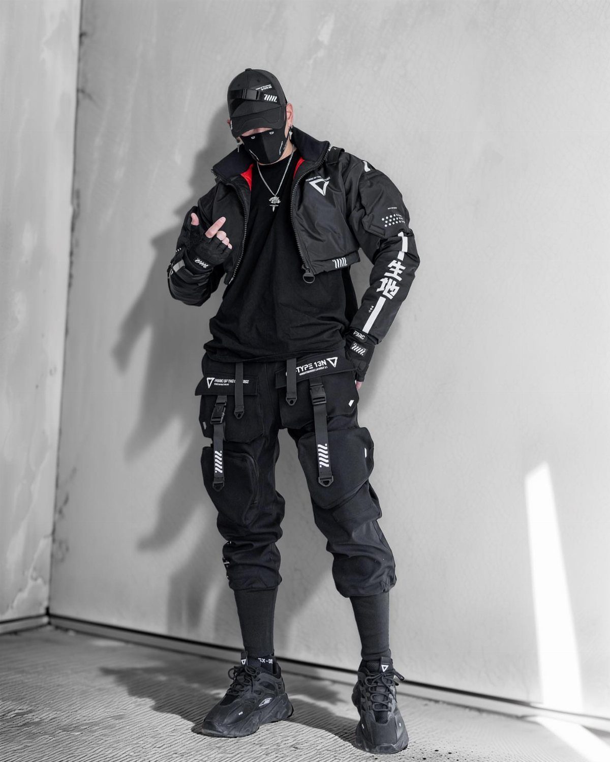A person in black techwear clothing including cargo pants and a jacket standing against a wall.