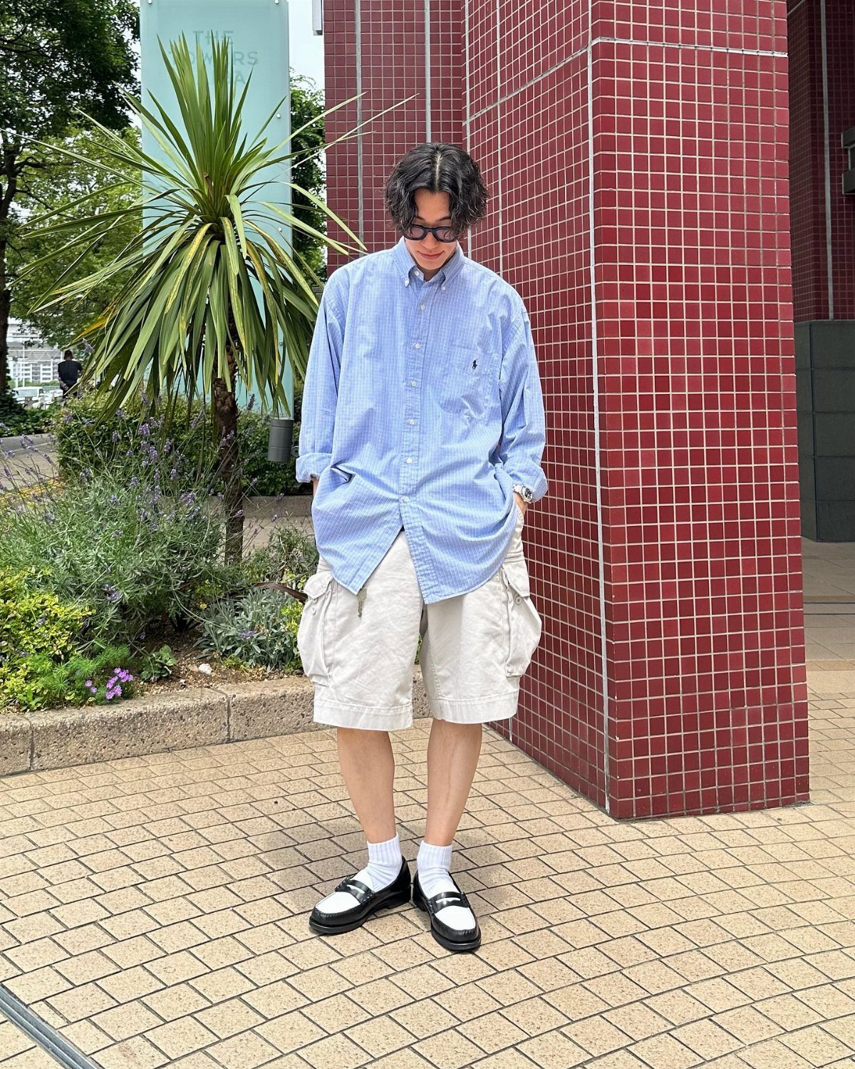 A person wearing a blue striped shirt, beige cargo shorts, and black loafers standing outdoors.