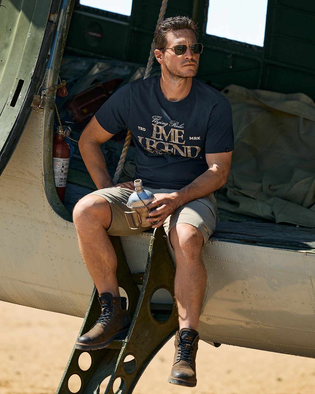 Man in cargo shorts and boots sitting on an aircraft holding a metal water bottle.