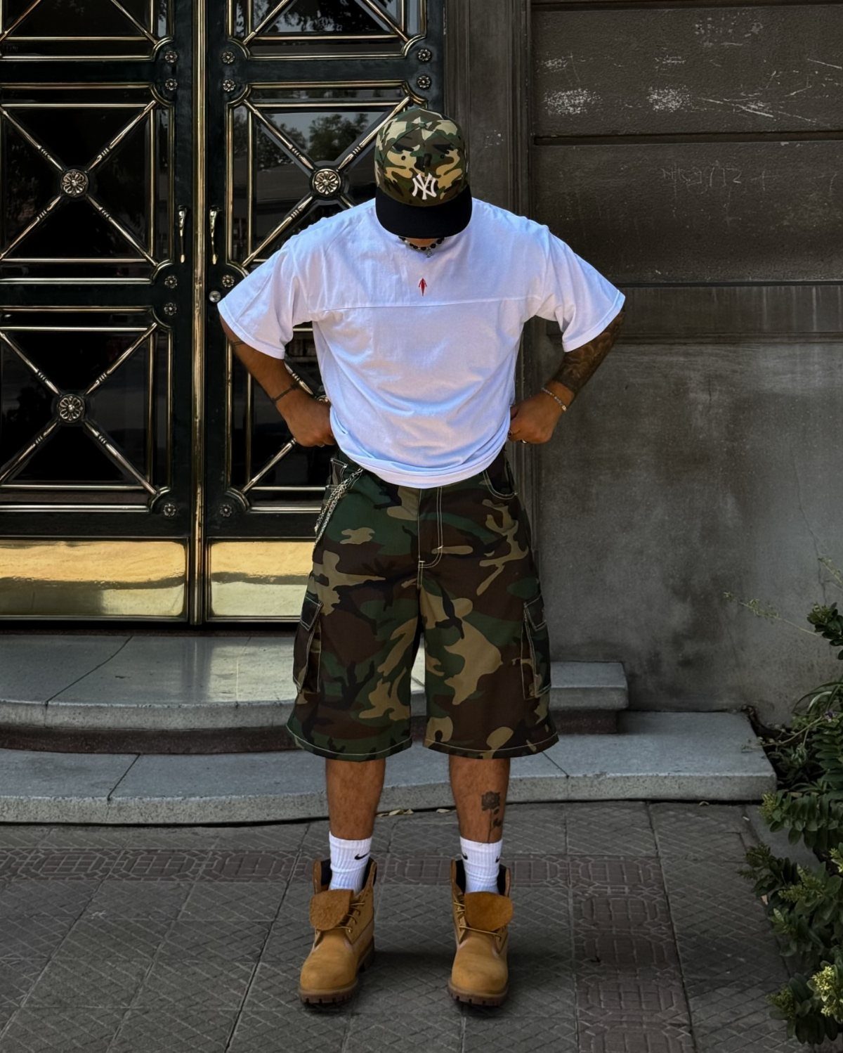 A person wearing a white oversized t-shirt and camouflage cargo shorts with tan boots.