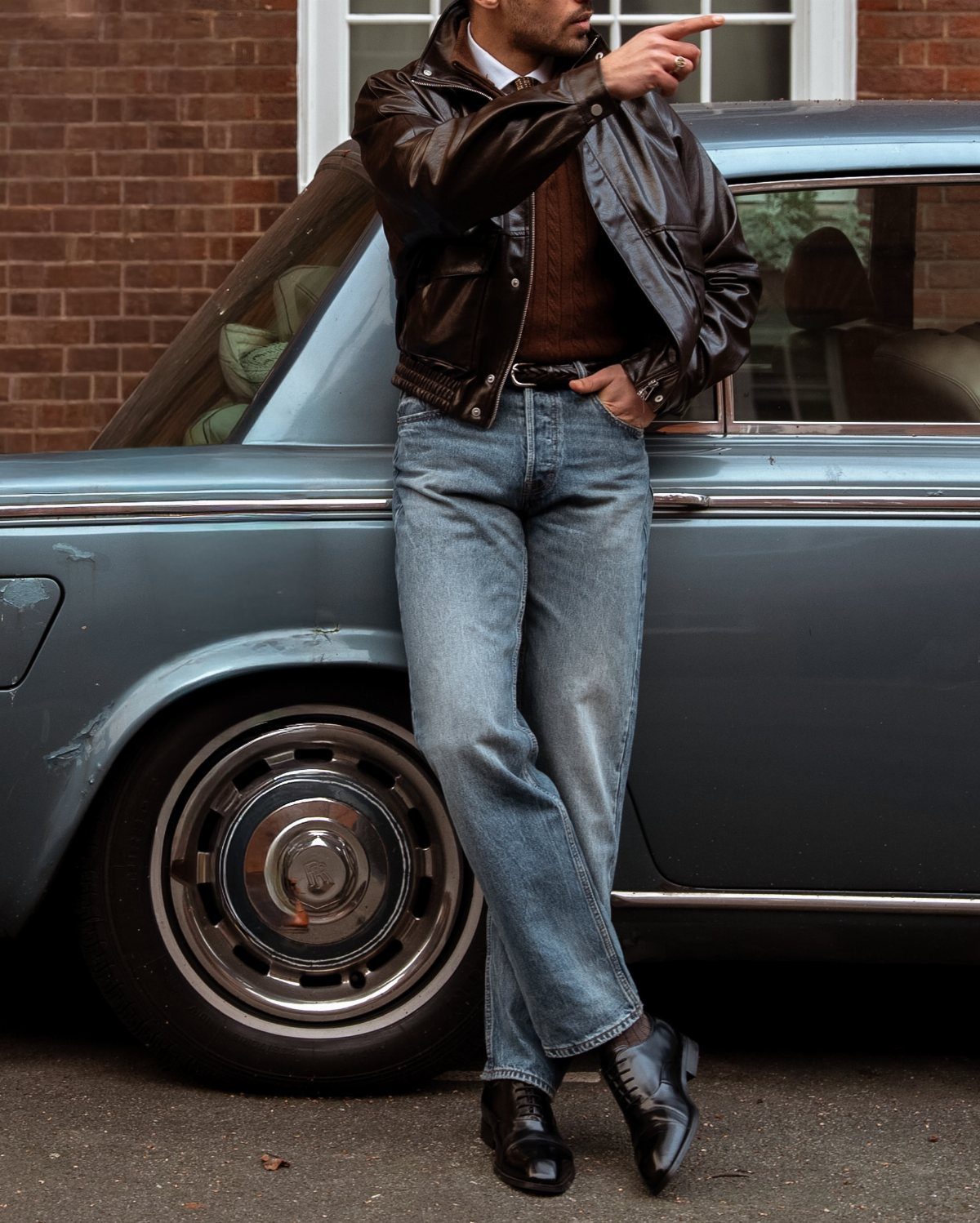 Man in leather jacket and blue jeans leaning against a vintage blue car wearing black loafers.