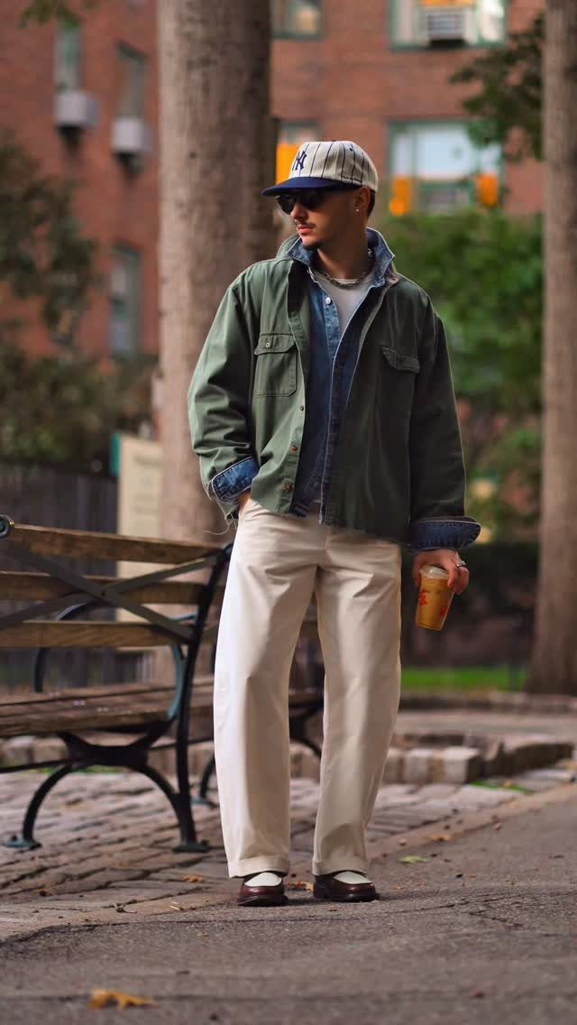 Man in a green utility jacket, denim shirt, and cream trousers standing in a city park.