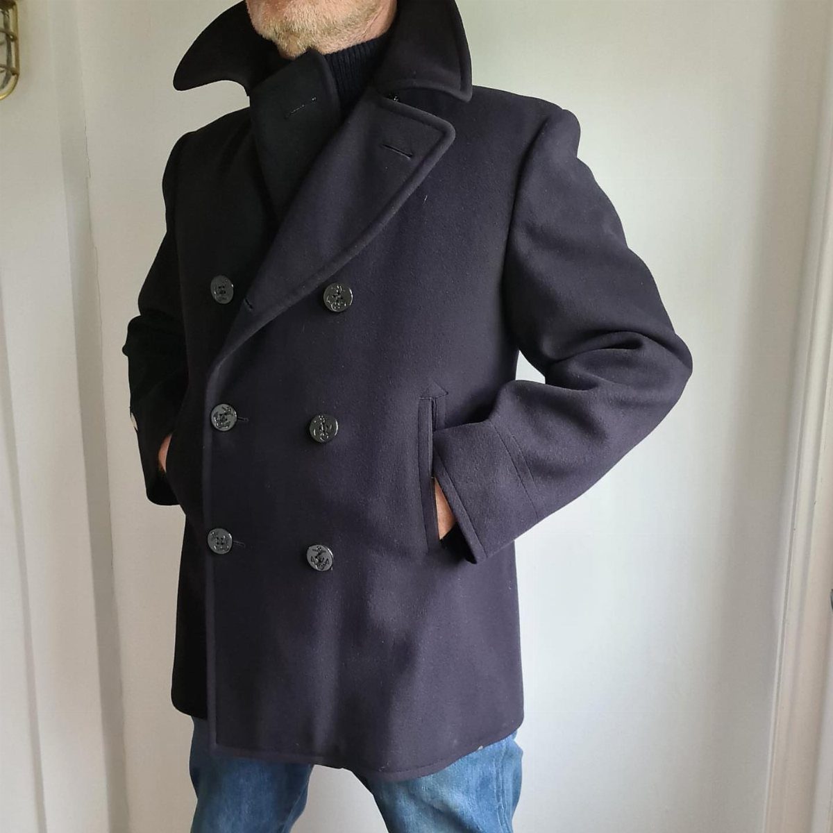 Man wearing a navy pea coat over a black turtleneck sweater and blue jeans.