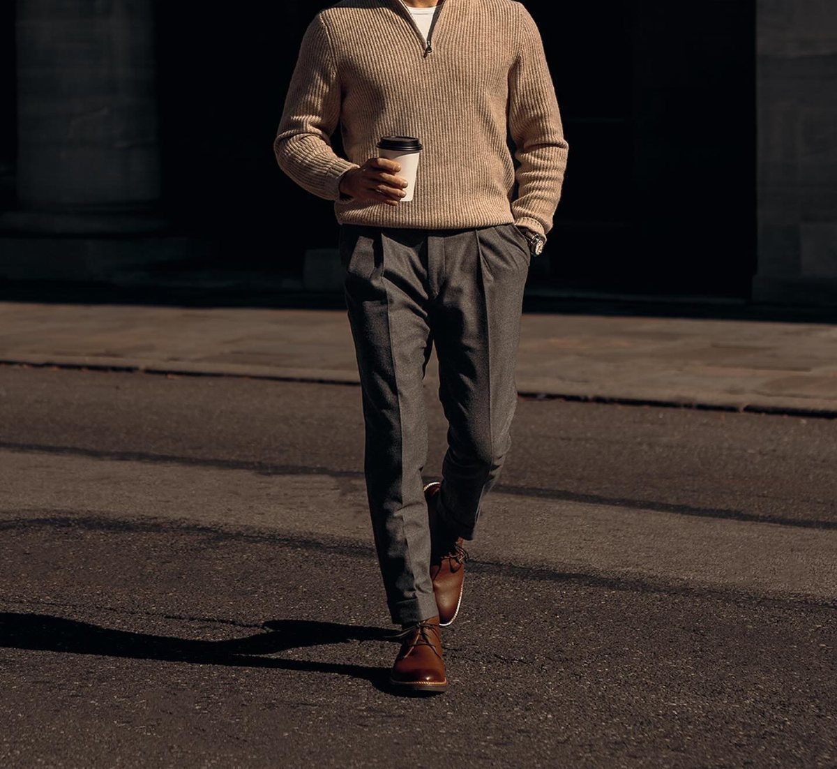 Man walking on a city street wearing a beige knitted sweater and grey pleated trousers.