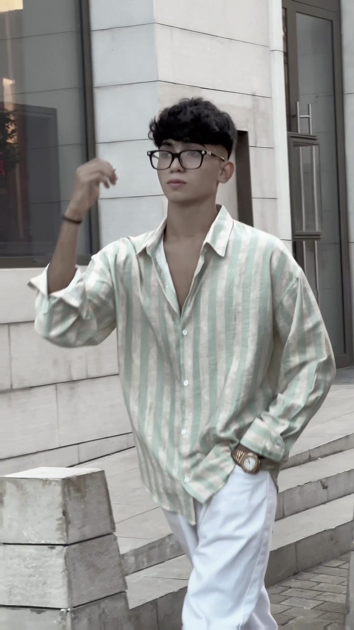 A man wearing a light green striped poplin shirt and white trousers standing outdoors.