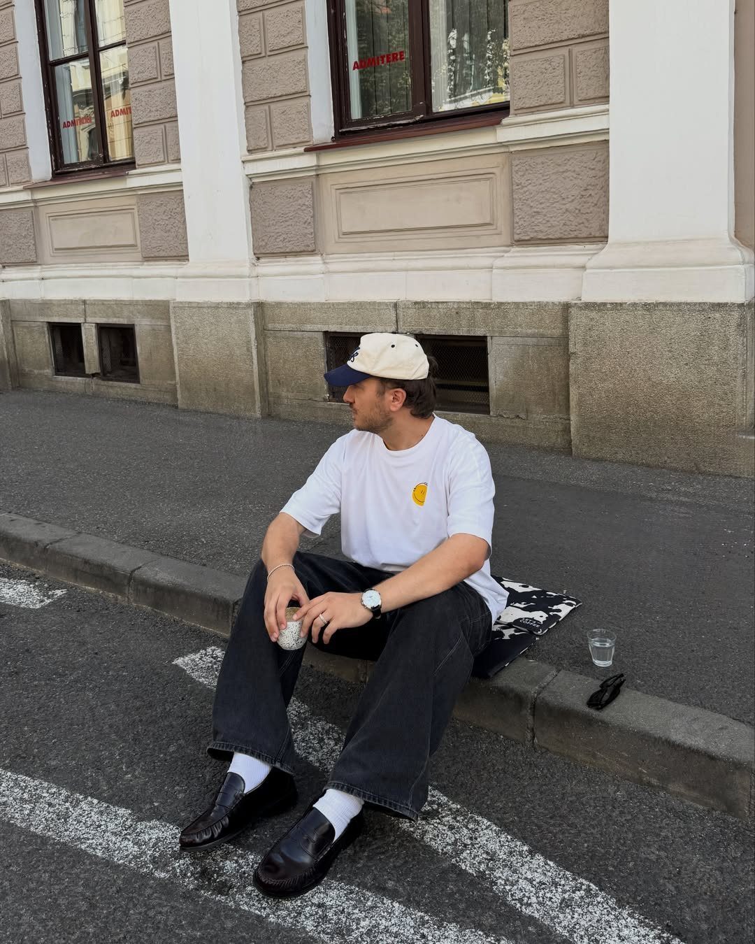 Man in white t-shirt and dark trousers sitting on a curb wearing black leather loafers.