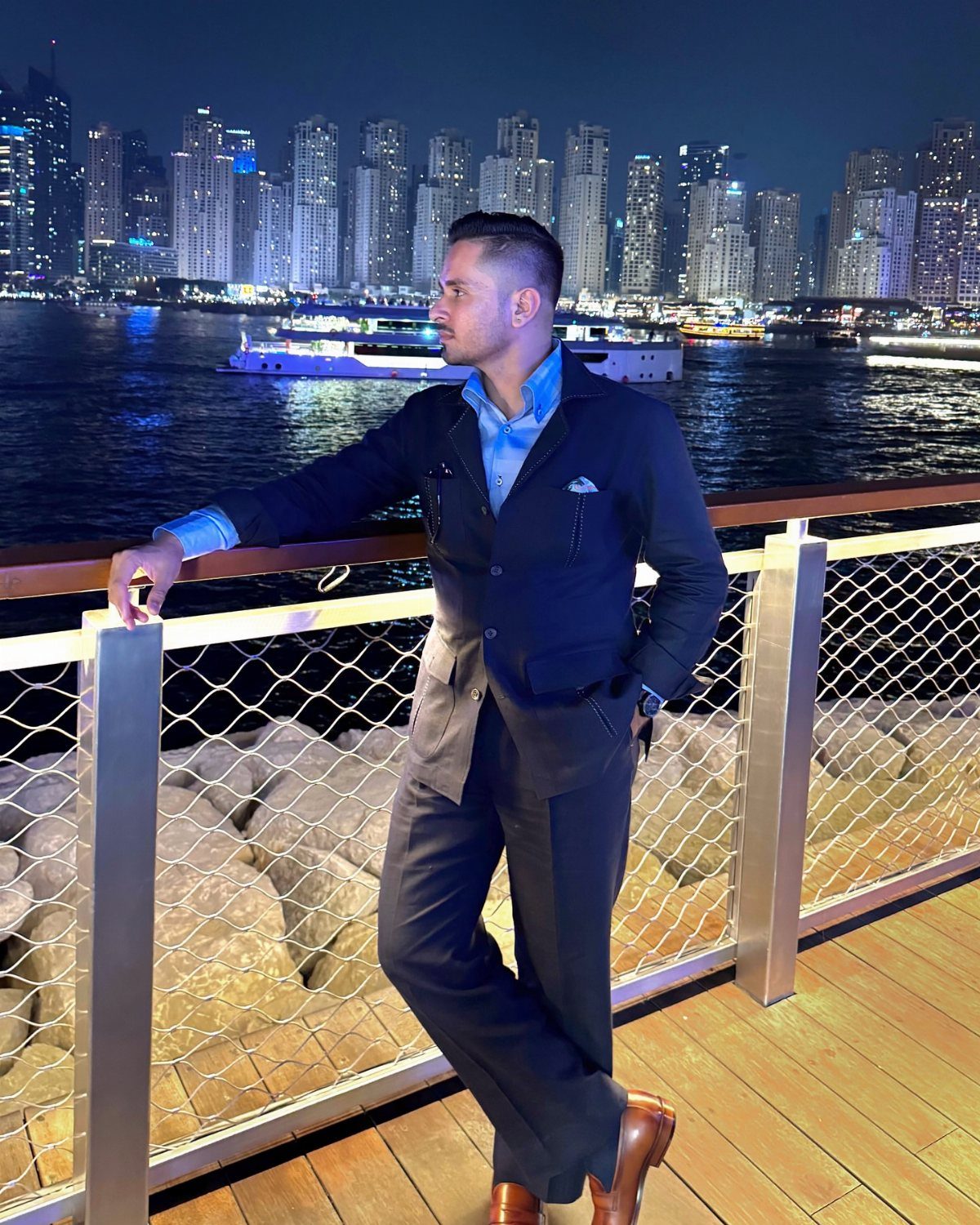 A man in a navy tailored suit posing by a waterfront railing against a city skyline.