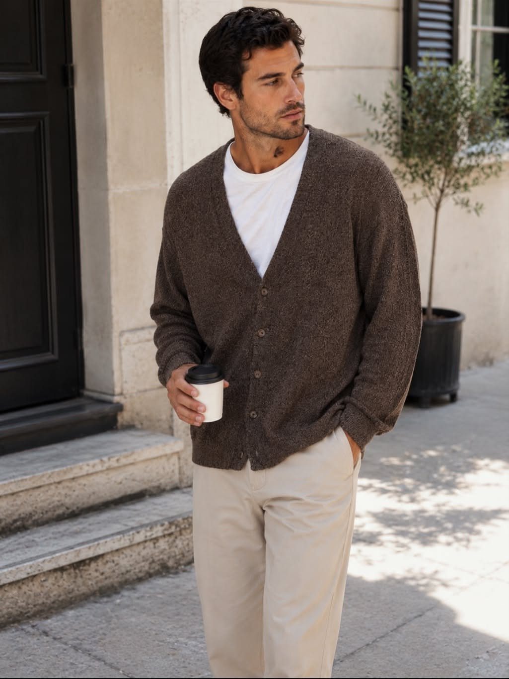 A man wearing a brown knit cardigan and beige trousers holding a coffee cup outdoors.