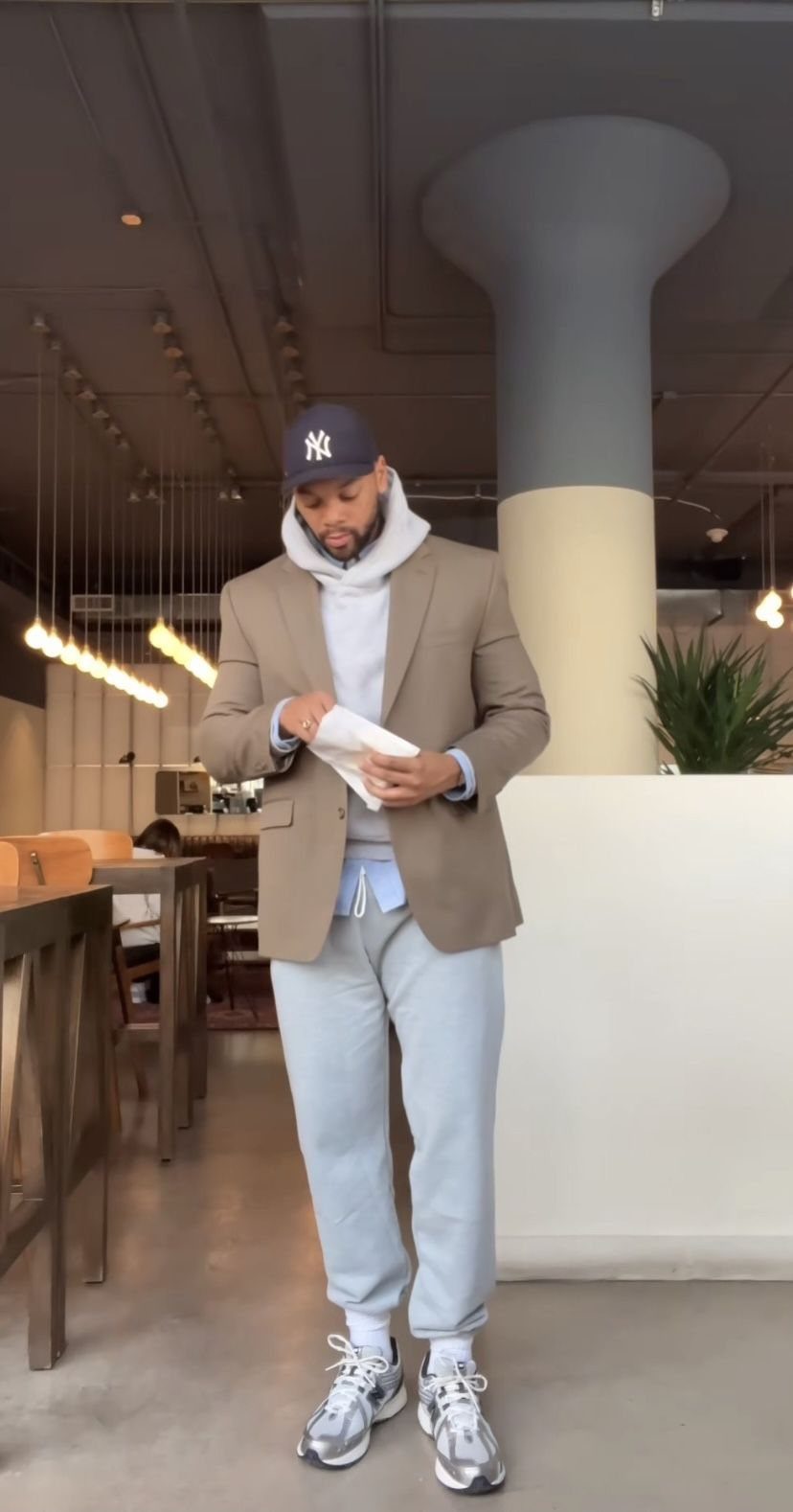 Man wearing a tan blazer, white hoodie, light blue joggers, and white sneakers indoors.
