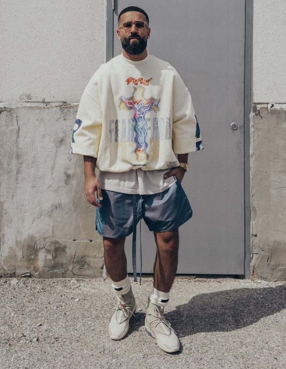 Man in an oversized graphic sweatshirt, blue shorts, and beige sneakers standing against a grey door.