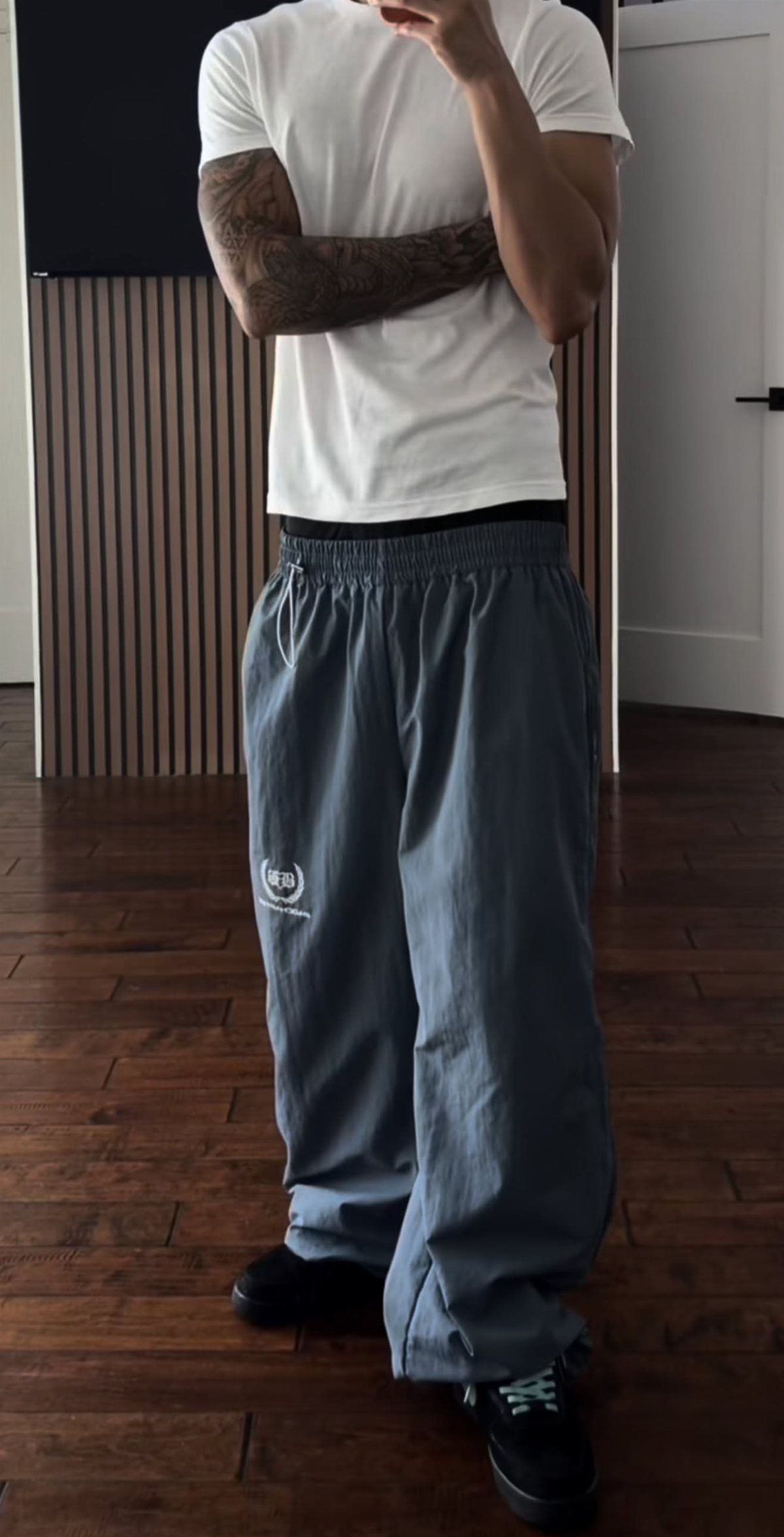 Person wearing a white t-shirt and grey track pants standing on a wooden floor.