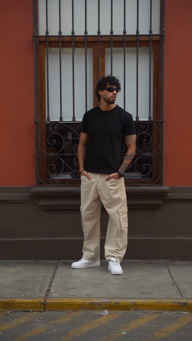 A man in a black t-shirt and beige cargo pants standing outdoors by a window.