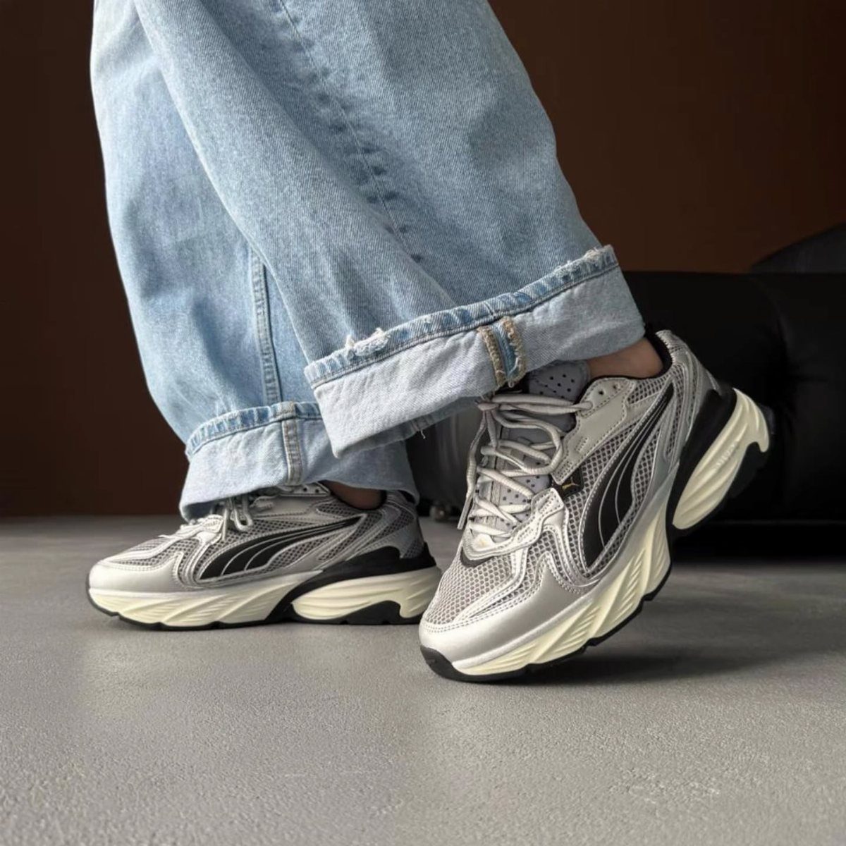 Silver and black chunky sneakers worn with light blue denim jeans on a grey floor.