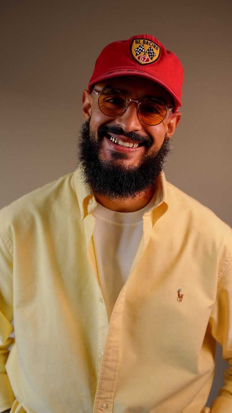 A bearded man smiling while wearing a red cap, tinted glasses, and a yellow button-down shirt.