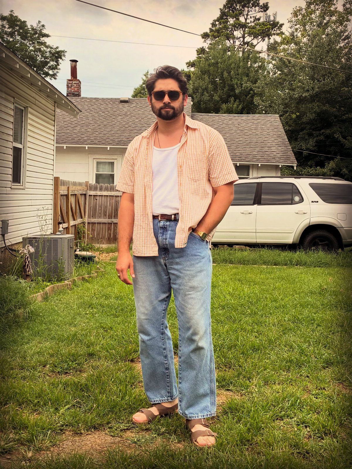 A man standing in a yard wearing a peach striped shirt and relaxed-fit denim jeans.