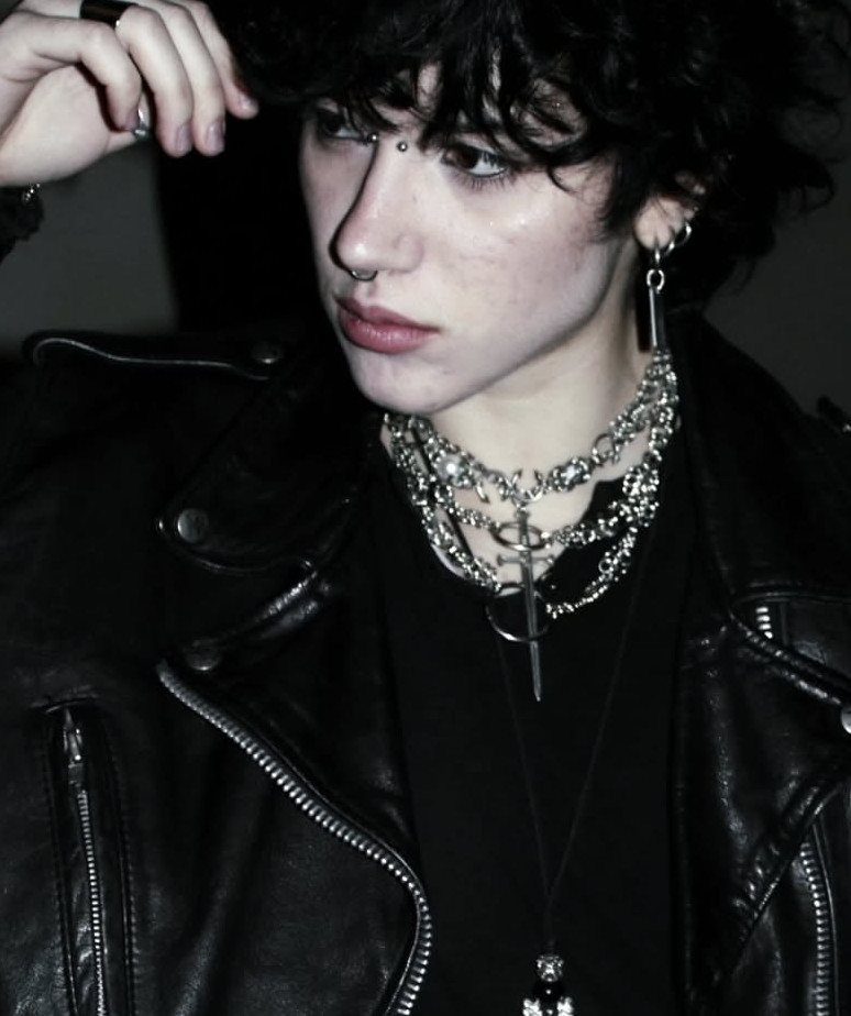 Person with dark curly hair wearing a black leather jacket and layered silver necklaces.