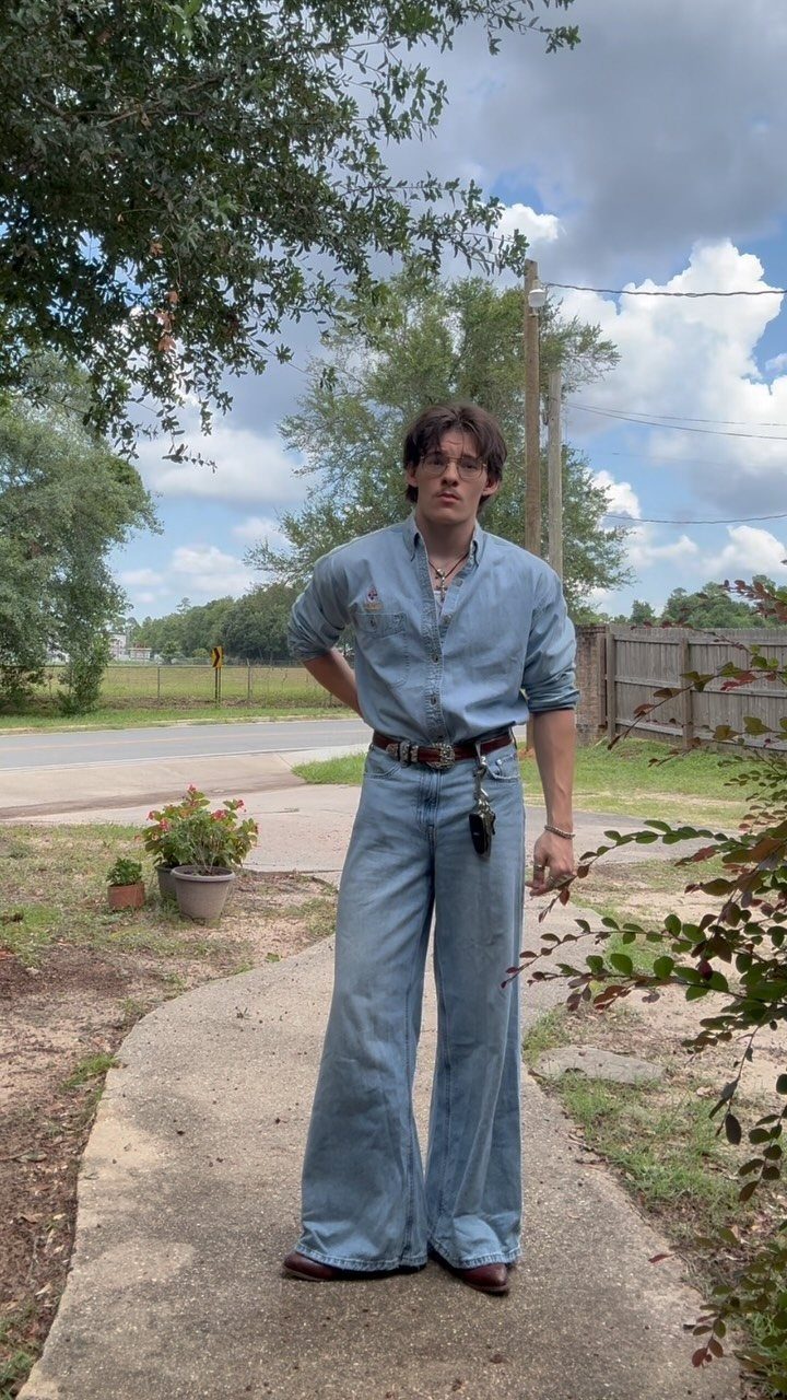 A young man wearing a denim shirt and wide-leg denim jeans standing on a path.