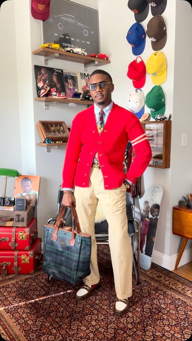 Man in a red cardigan, patterned shirt, and tan trousers holding a plaid tote bag.