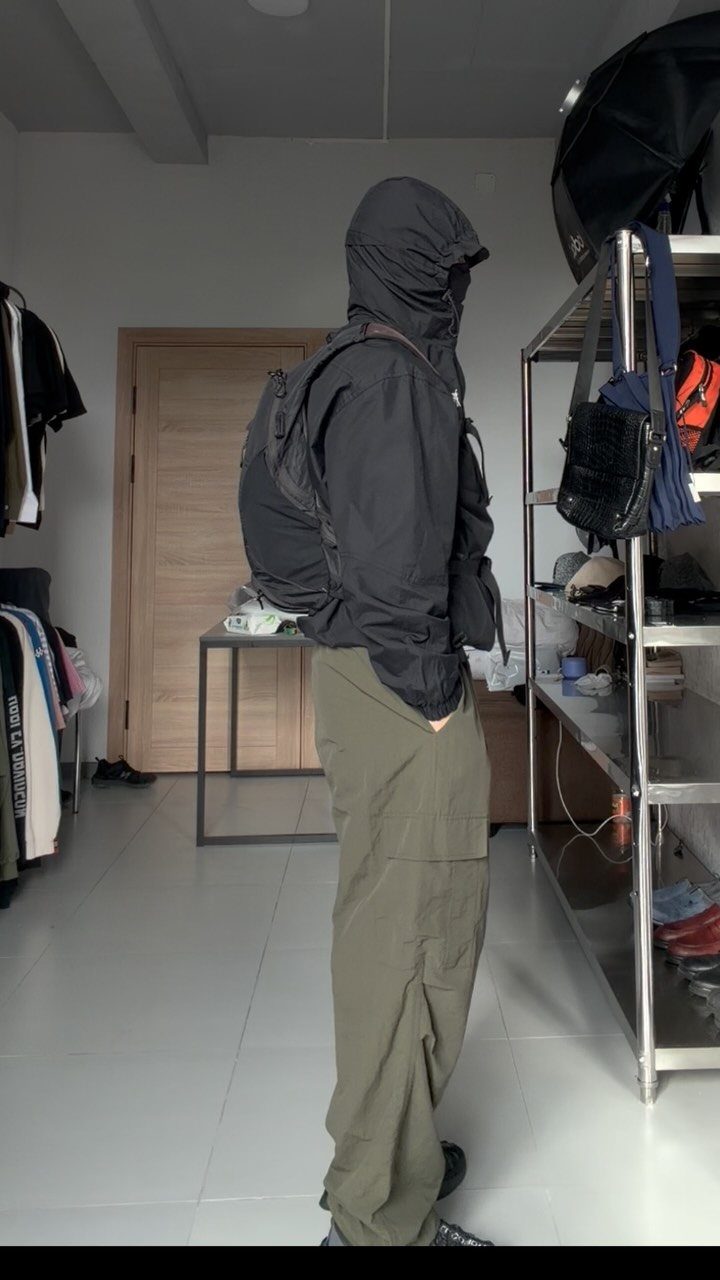A person wearing a black technical hooded jacket, olive cargo pants, and a black backpack.
