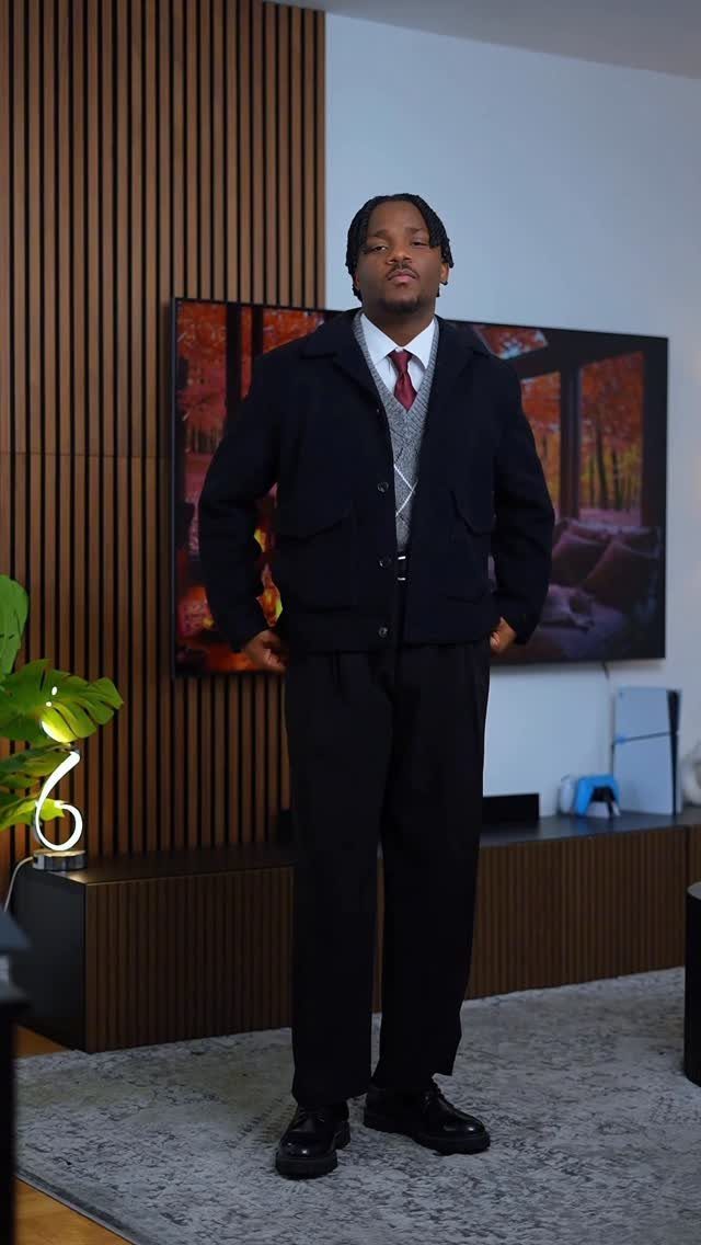A man wearing a navy jacket, grey sweater vest, and red tie standing indoors.