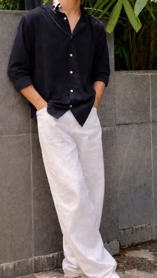 Man wearing a black button-down shirt and wide-leg white linen trousers leaning against a wall.