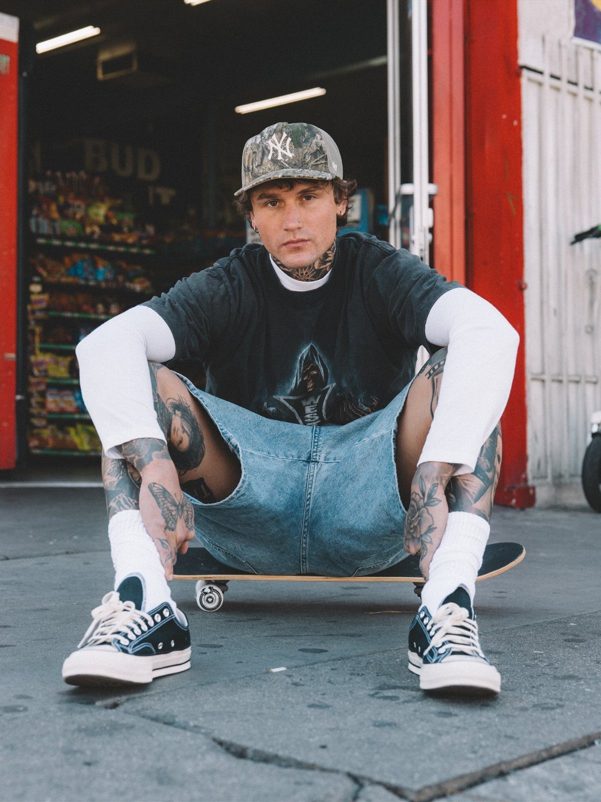 A tattooed man in streetwear sitting on a skateboard outdoors in front of a store.