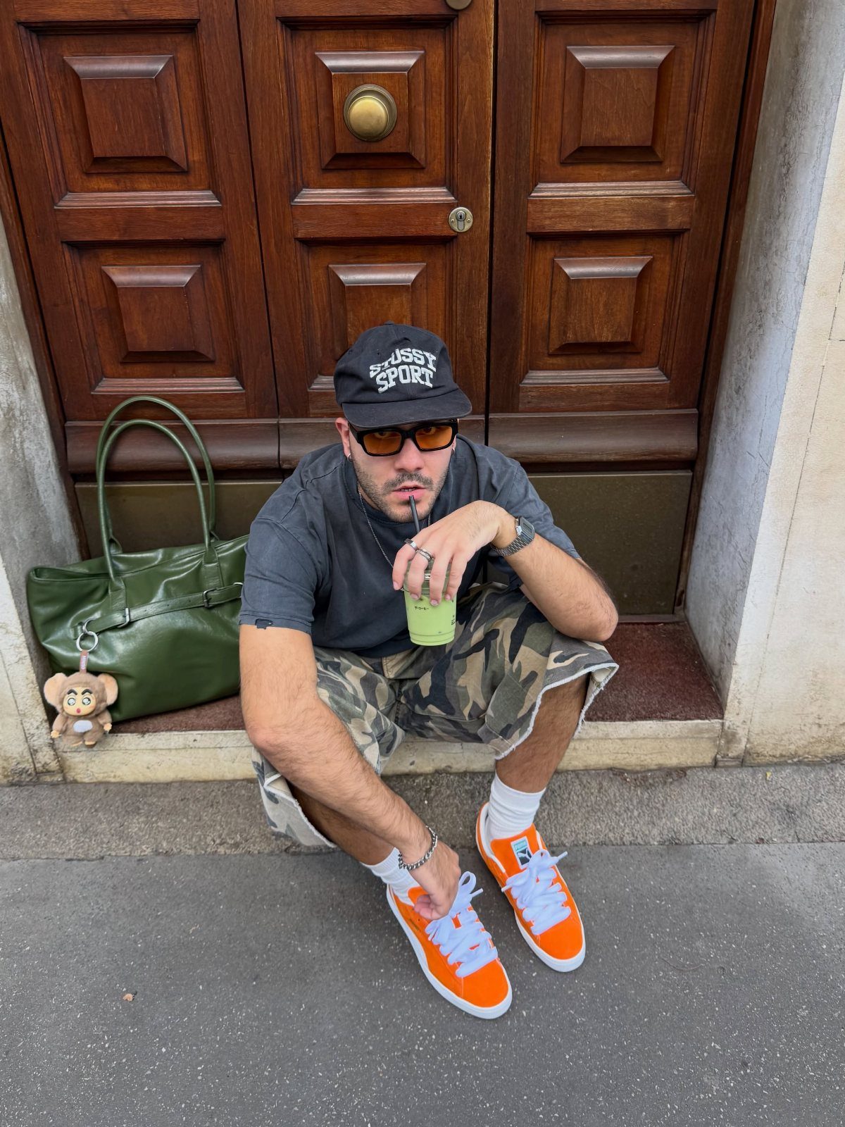 Man in camouflage shorts and orange sneakers sitting in front of a wooden door.