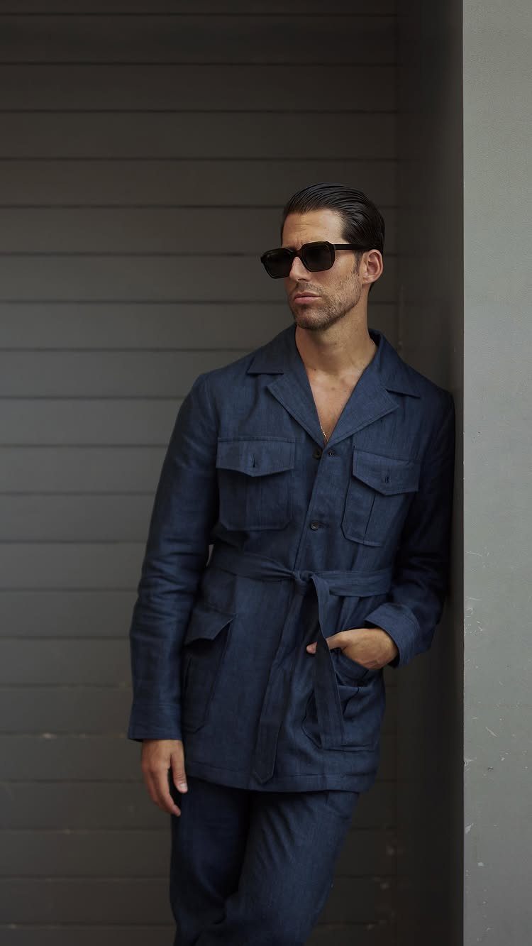Man in a navy blue belted utility jacket and sunglasses leaning against a grey wall.